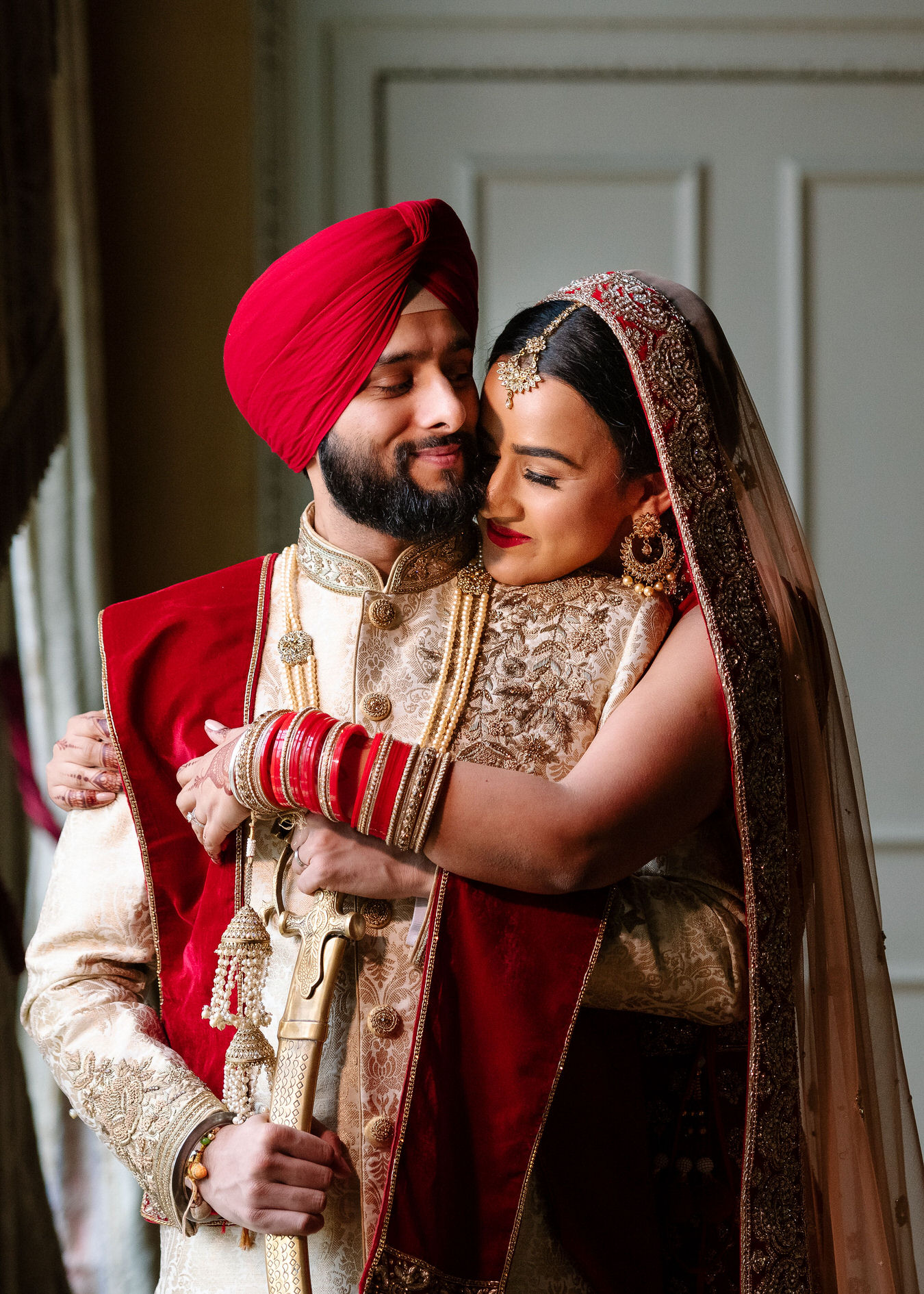 Bride is hugging with a hand the groom from the back laying her chin on his shoulder. Groom has his kirpan in his hand