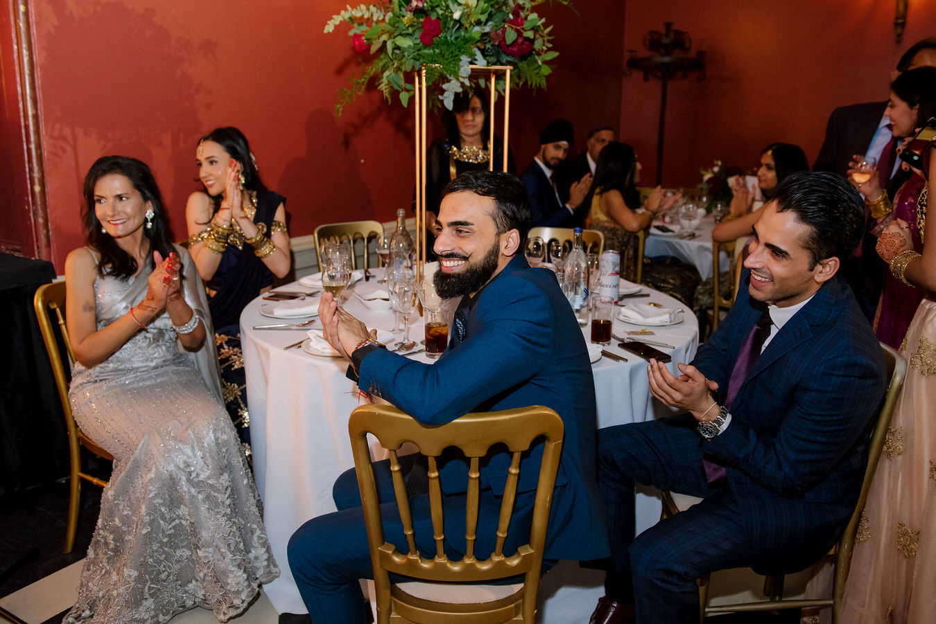 Indian wedding guests at a table near the bride and groom are applauding the speeches.