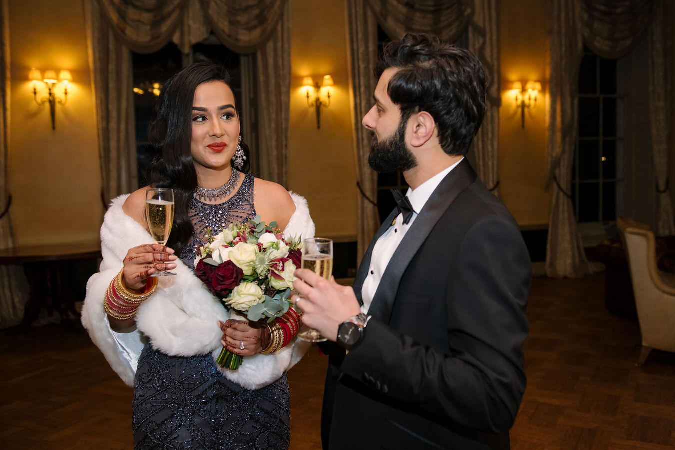 Asian bride dressed in a grey sparkling evening dress and the groom with champagne glasses in their hands are looking at each other