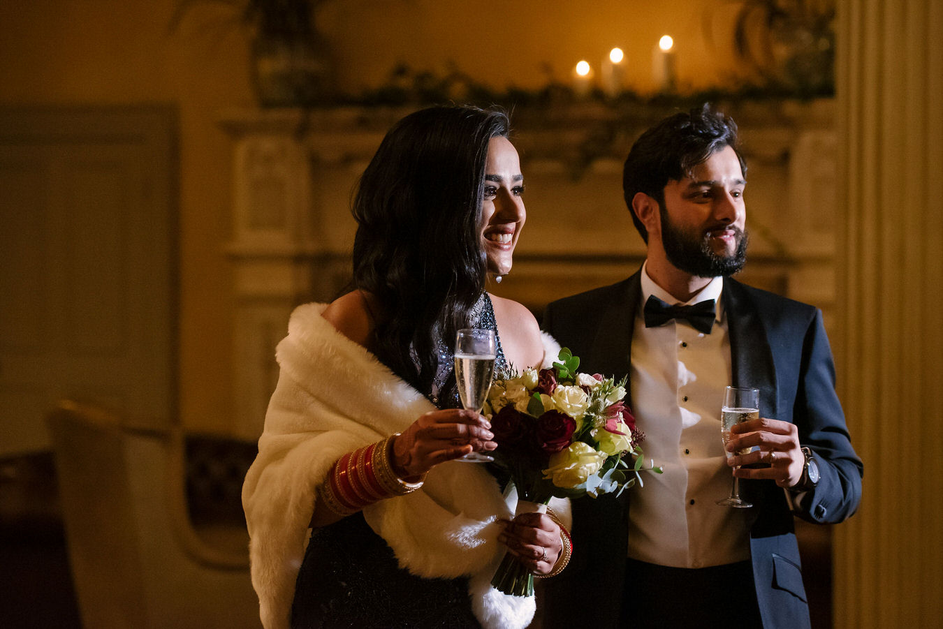 Asian bride with a white fur shawl and groom with black smoking suit, having champagne glasses in hands are having fun.