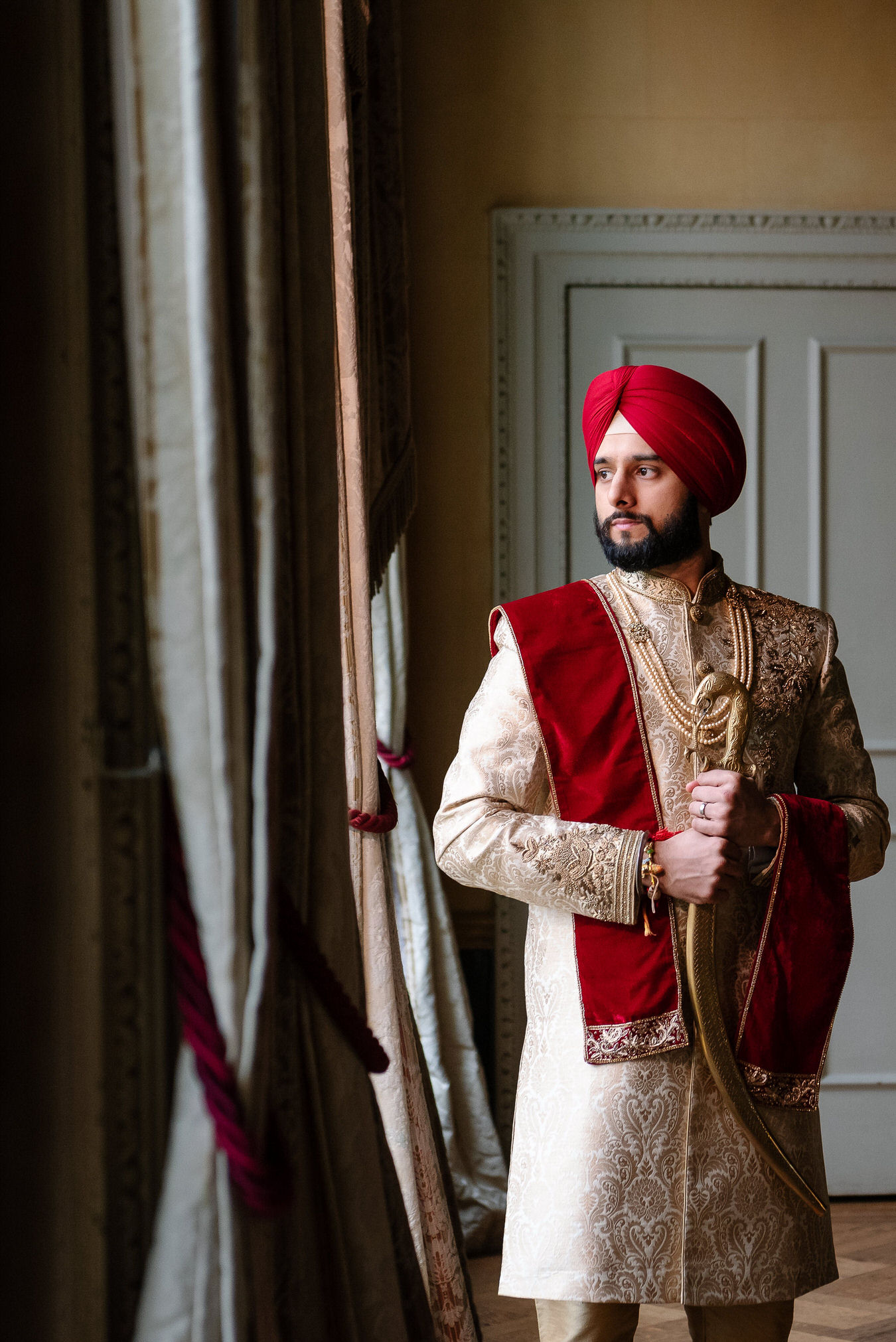 Sikh wedding groom with kirpan in his hands and a red turban on the head looking at his right.