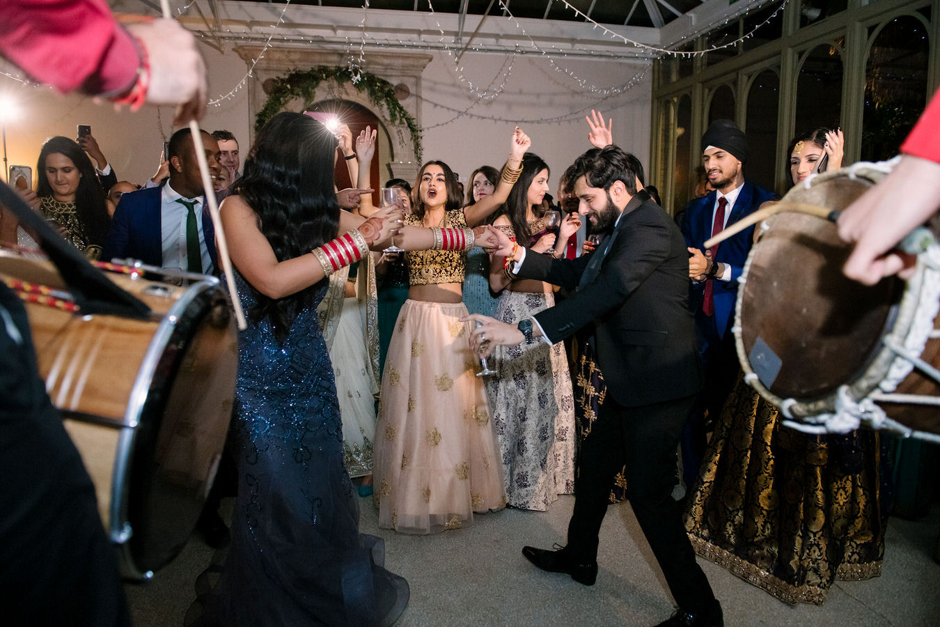 Asian wedding couple dancing with glasses in their hands along with other guests. Asian Dhol drummers are playing in the foreground.