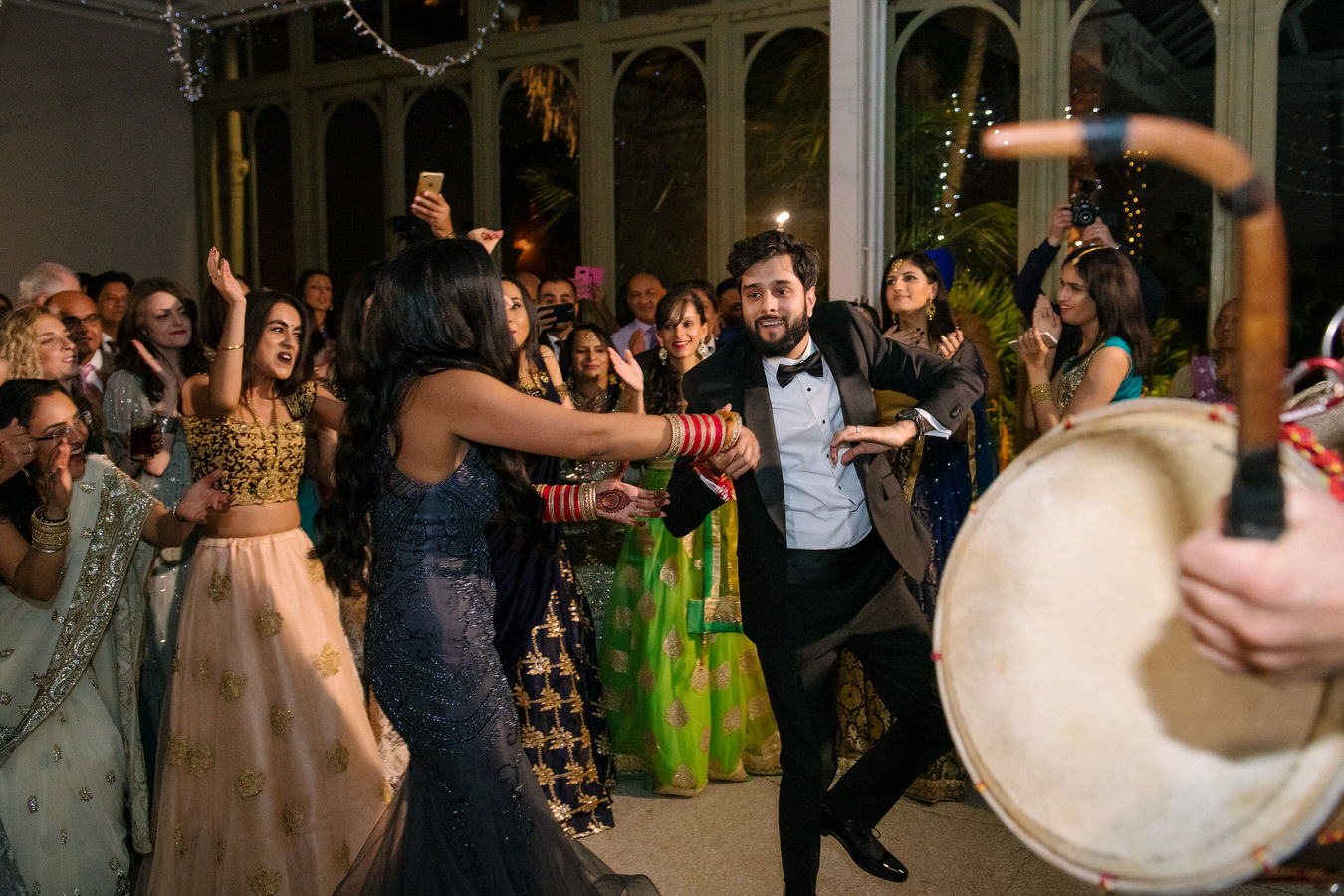 Sikh Asian wedding couple are dancing in the middle of the dancefloor and other guests applaud around. In the foreground there is a Dhol Drum.