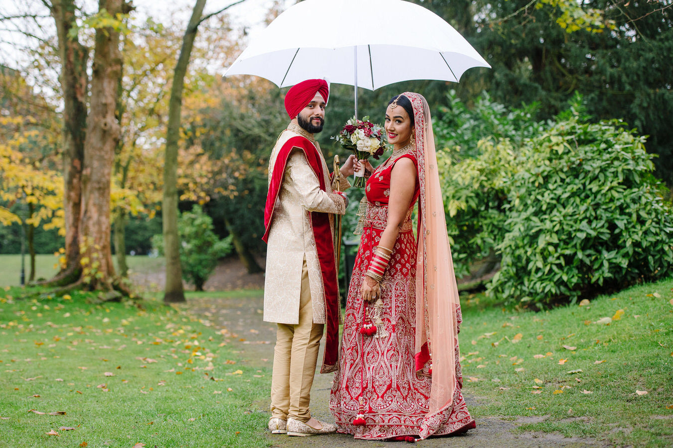 Sikh Asian wedding bride and groom in the garden holding a white umbrella and a bouquet in hands
