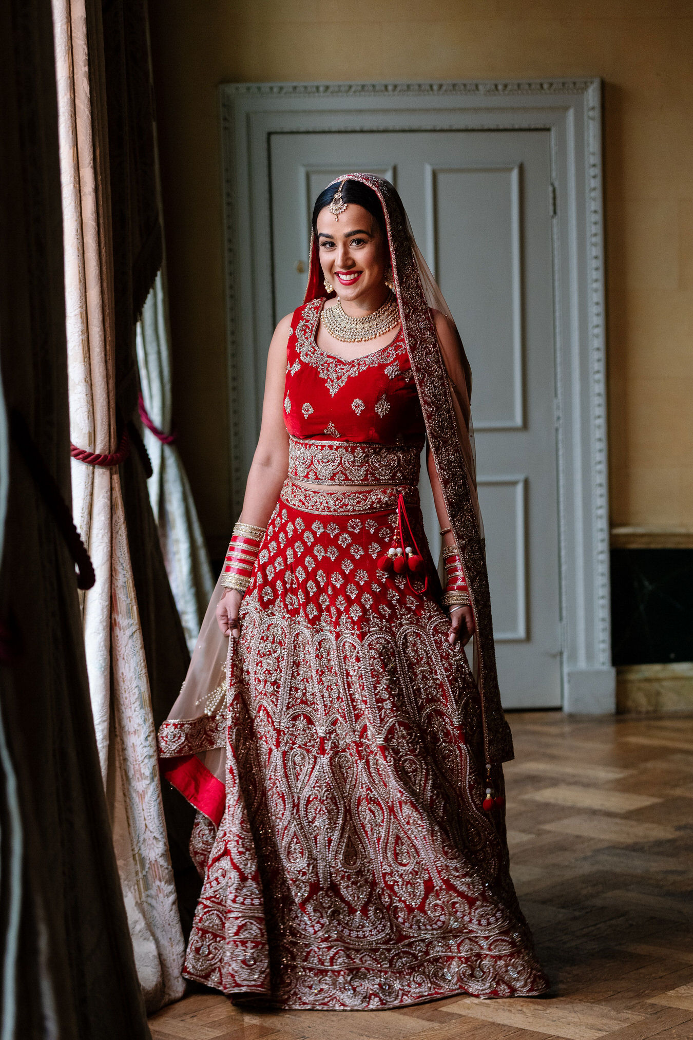 Asian Sikh bride dressed in a red bridal lehenga with golden details smiling in natural light.