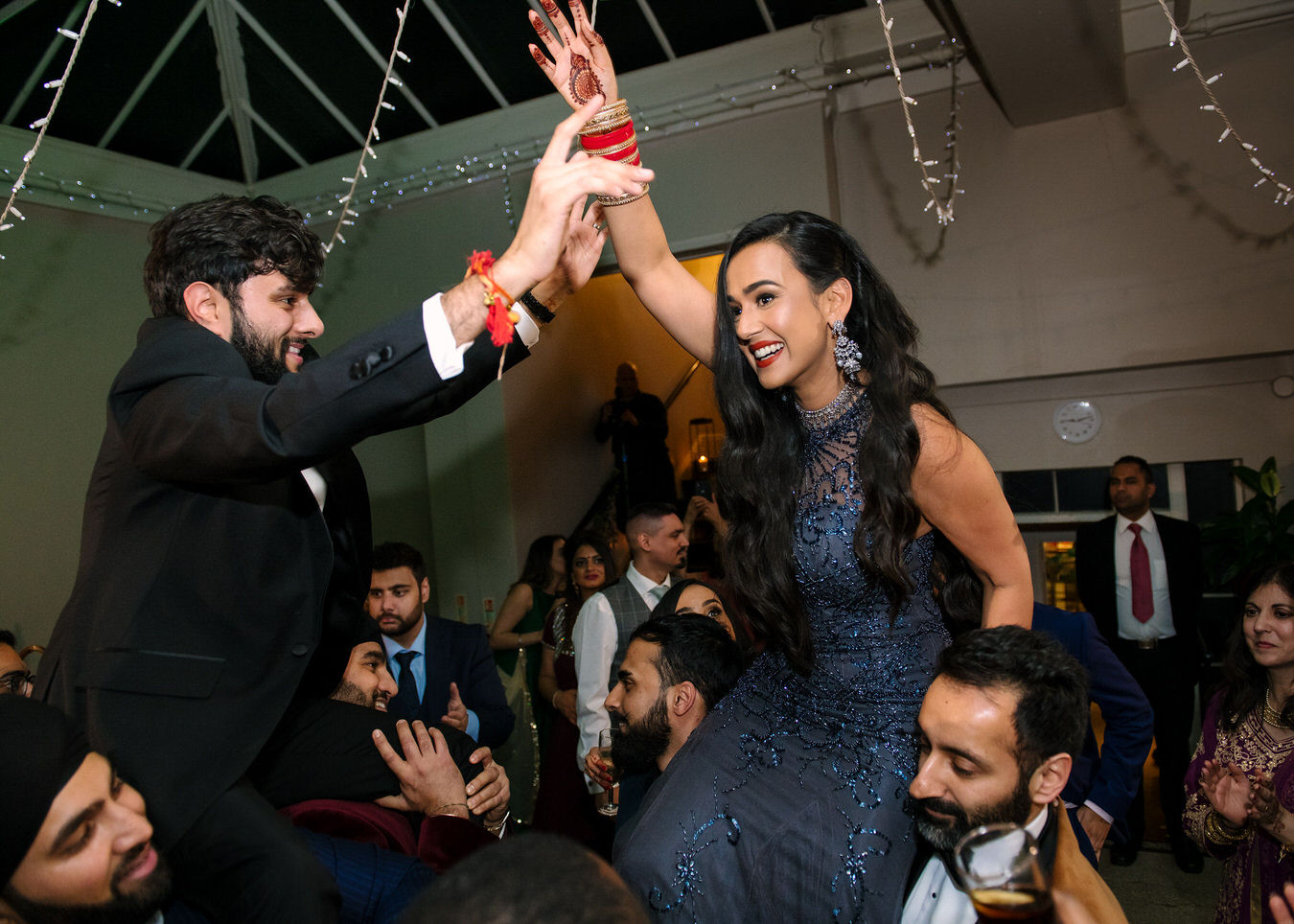 Sikh Asian bride and groom with hands in the air, singing and dancing on the guests shoulders.