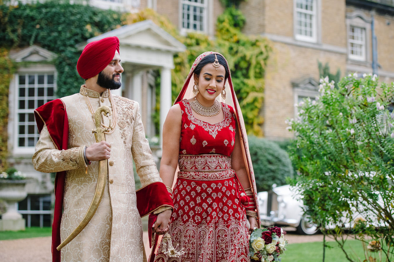 Sikh bride in a bridal long lehenga with Sikh groom in a Punjabi outfit with his kirpan in right hand walking in front of Hampton Court 
