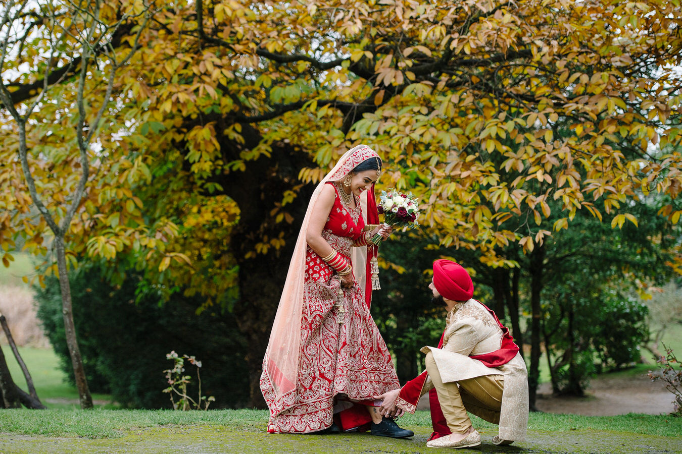 Asian groom tying bride’s black Converse shoes in the grounds at Hampton Court House looking autumnal as captured by Asian Sikh wedding photographer.