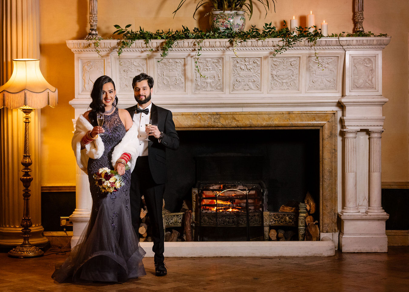 Asian bride in a grey long evening dress & white fur cape holding a red & white flower bouquet in her hand and groom dressed in a black smoking suit staying in front of the fireplace at Hampton Court