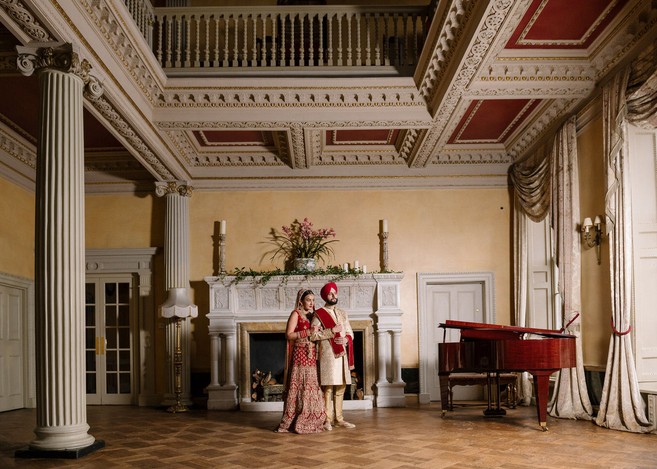 Sikh Asian wedding bride and groom close to each other in front of a white stone fireplace decorated with candles and flower pots at Hampton Court House